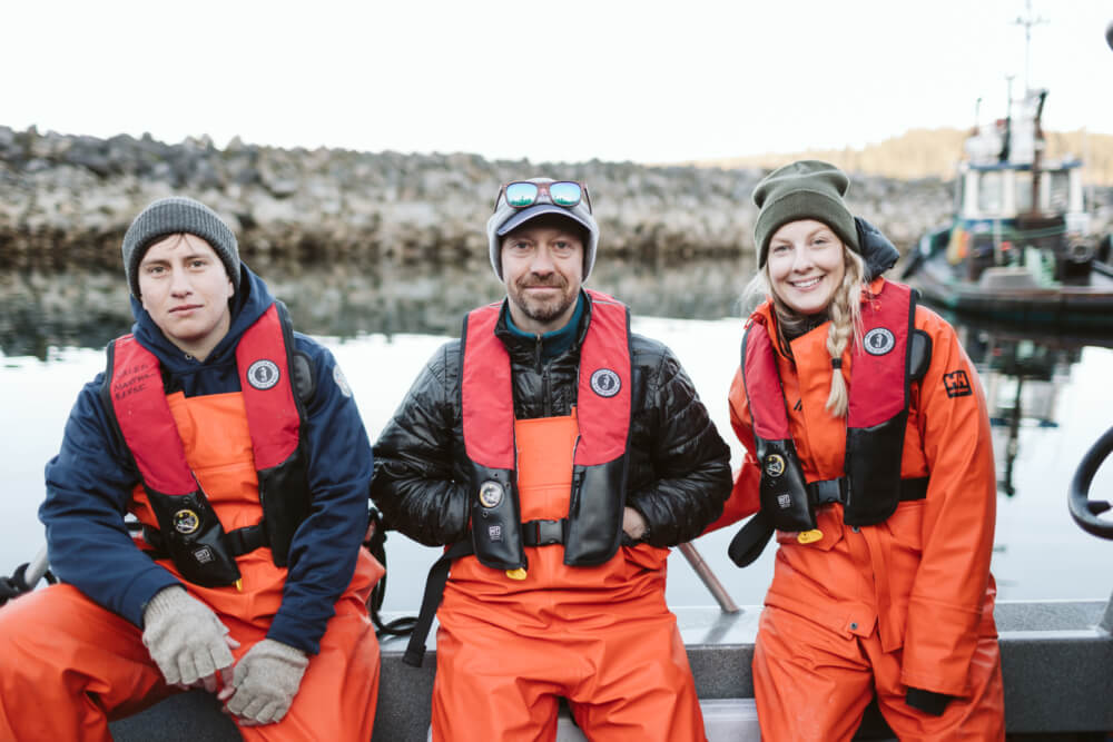 Three researchers sitting on a boat with orange hip-waders and lifejackets